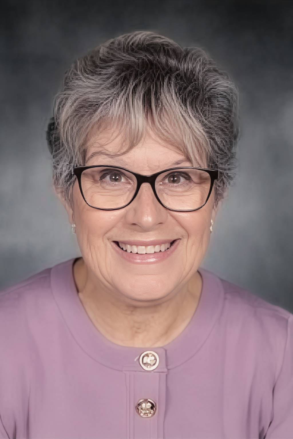 An older white woman with short grey hair smiles. She wears a mauve sweater and black framed cat's eye glasses